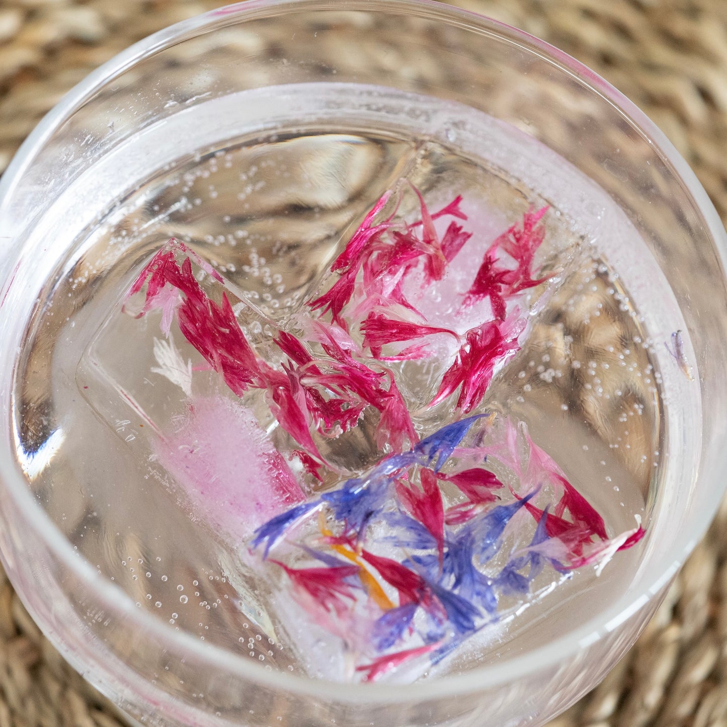 Clear glass with colourful flower petal ice cubes floating in water on a woven surface