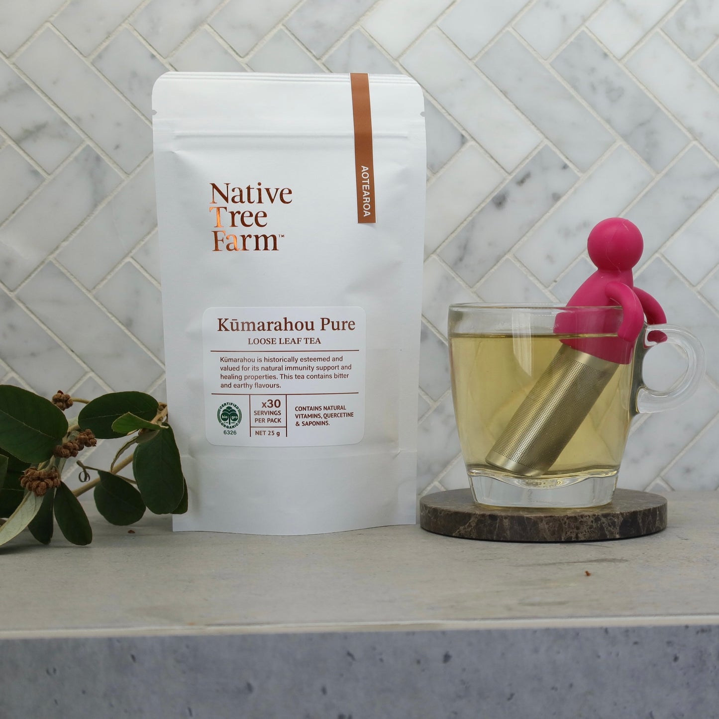 Native Tree Farm Kumarahou herbal tea pouch with a glass of tea and pink tea infuser on their marble kitchen bench