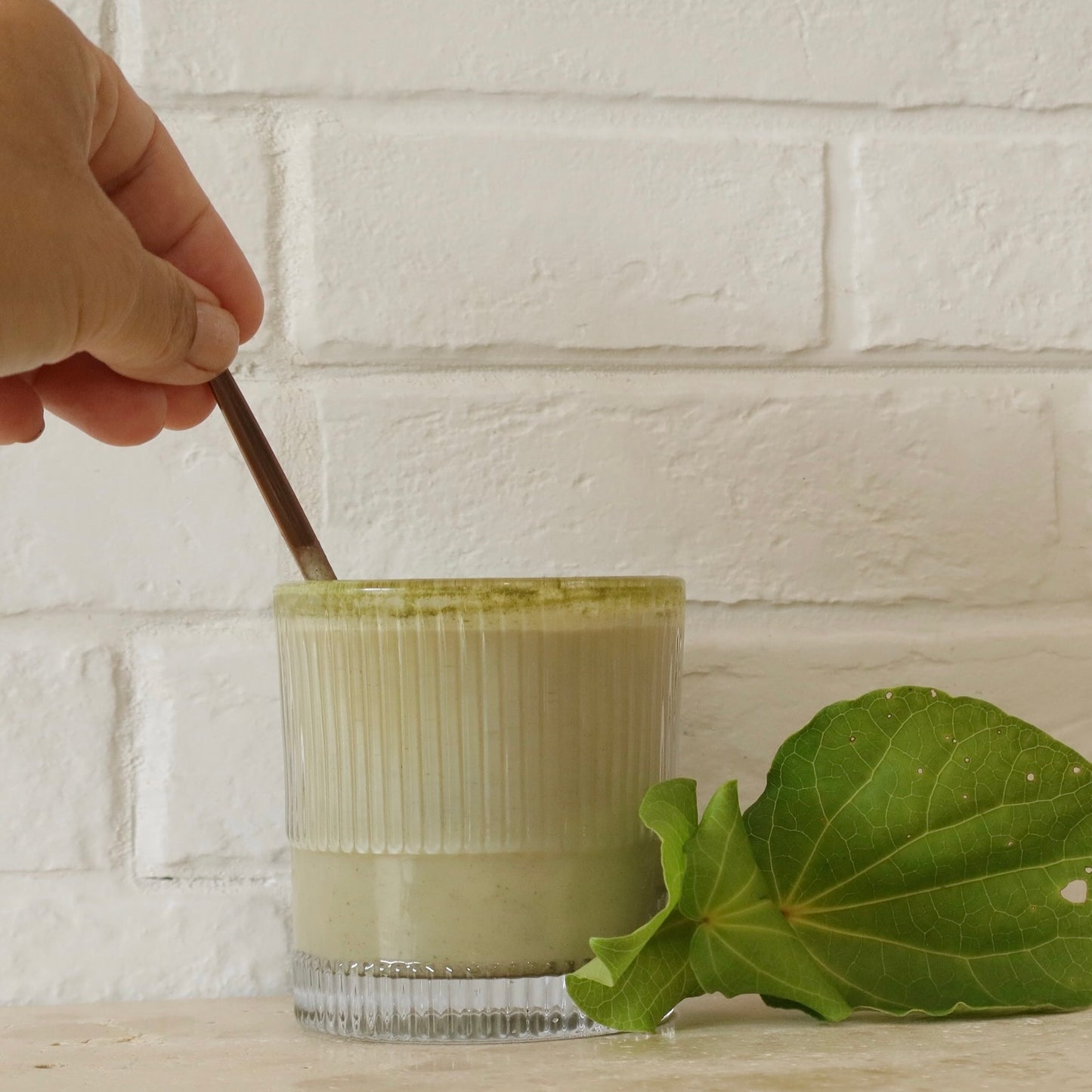 Hand stirring a green kawakawa latte in a glass with a leaf on a white brick wall background