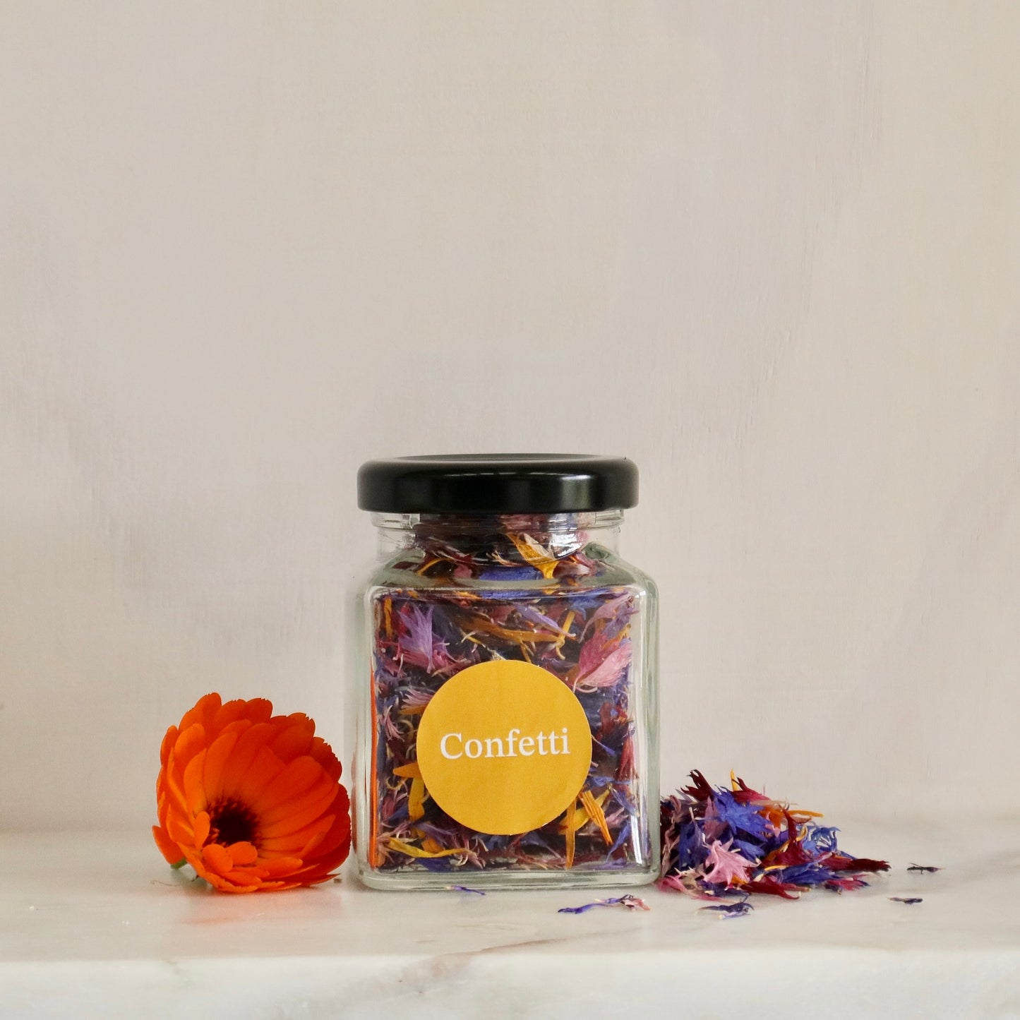 Native Tree Farm's Jar of confetti mixed flower petals with a yellow label on a white surface, featuring an orange flower.
