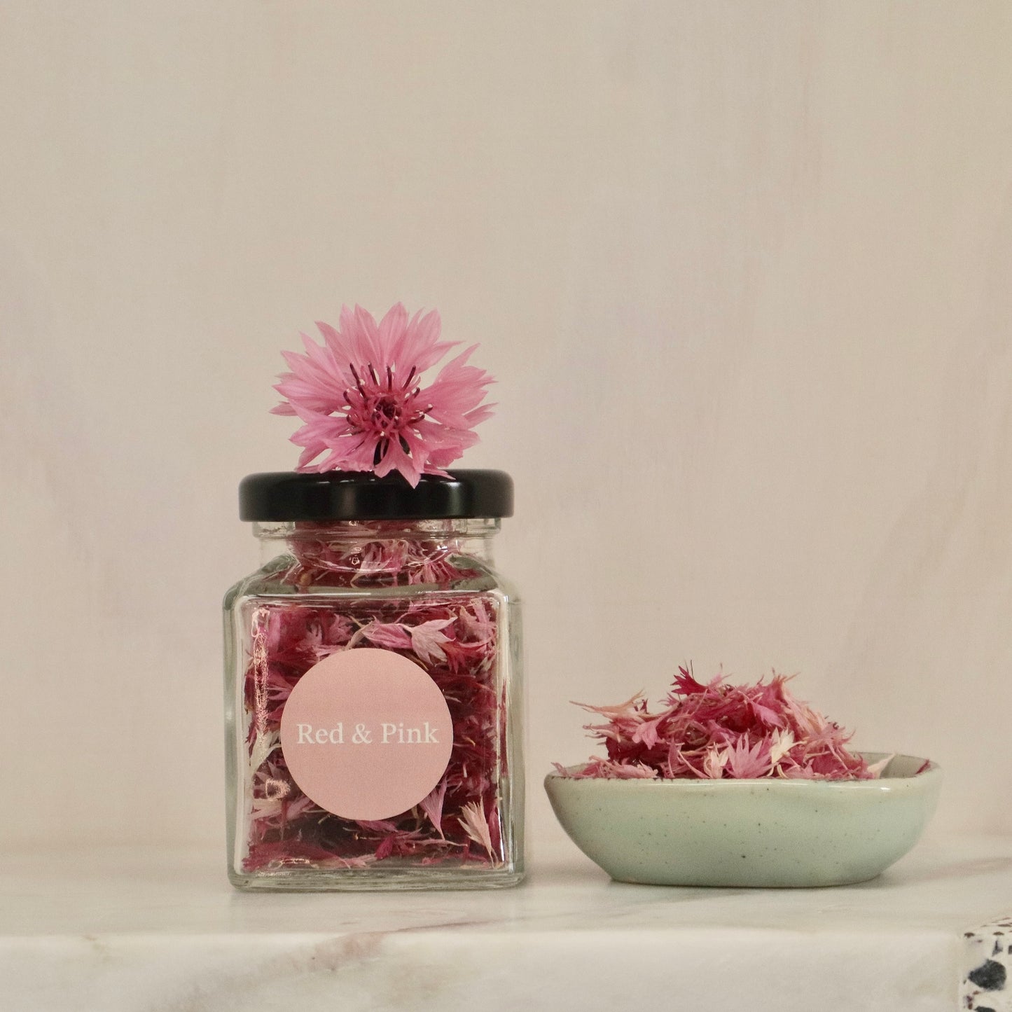 Jar of Native Tree Farm's pink organic edible flowers with a label and a bowl of flowers on a neutral background