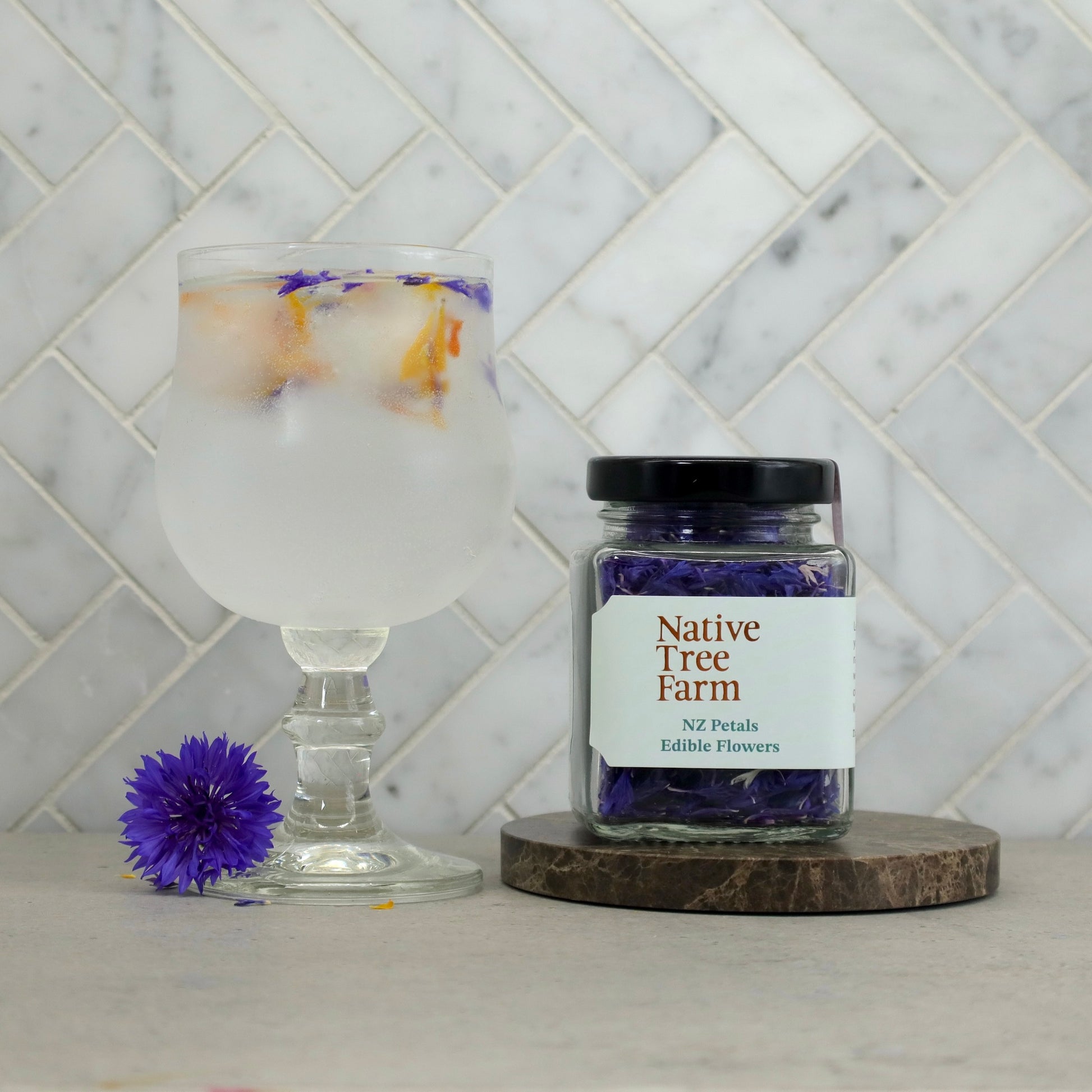 Jar of Native Tree Farm edible flowers on a stone table top with a glass drink in the background.