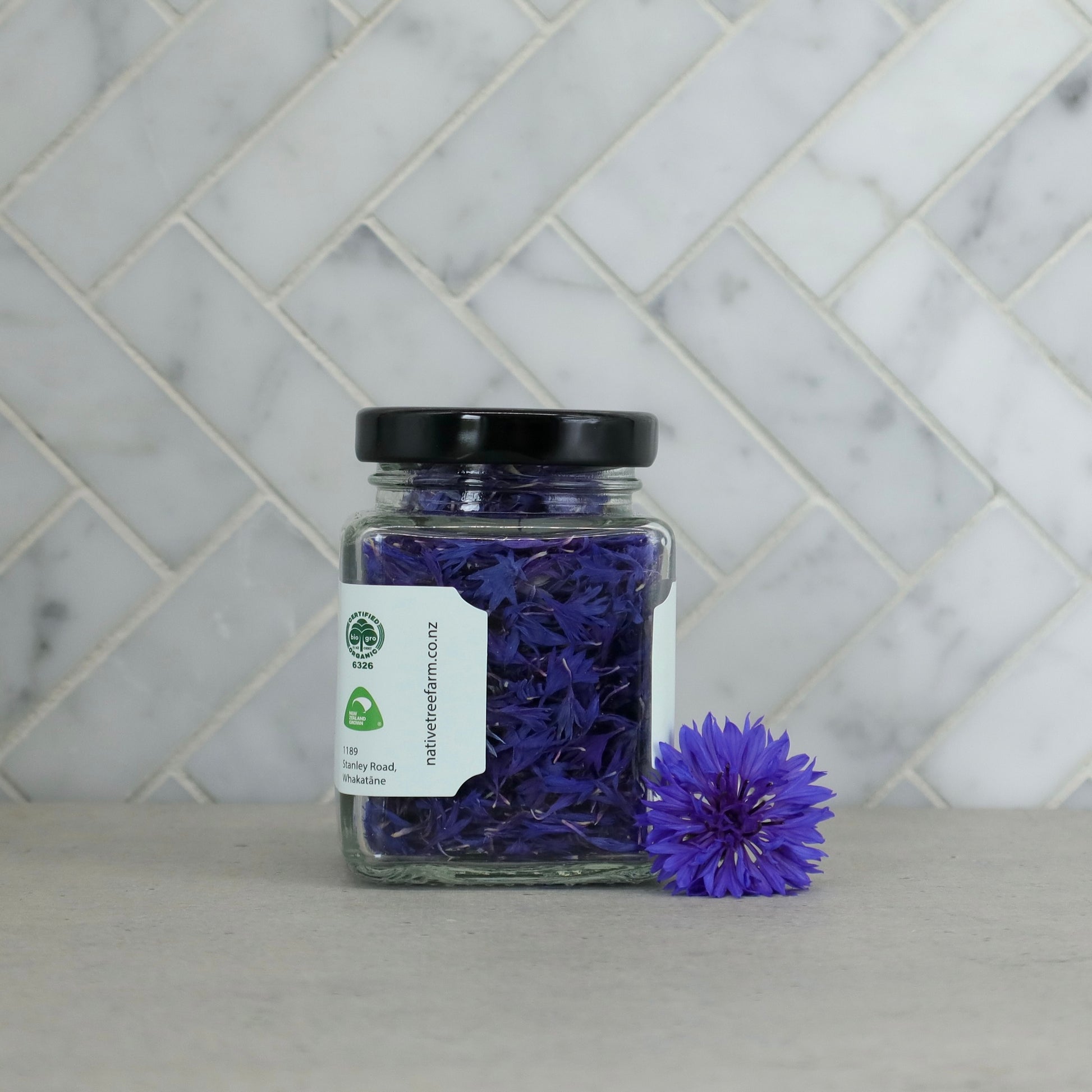 Jar of Native Tree Farm edible blue flowers next to a single blue flower on a marble table