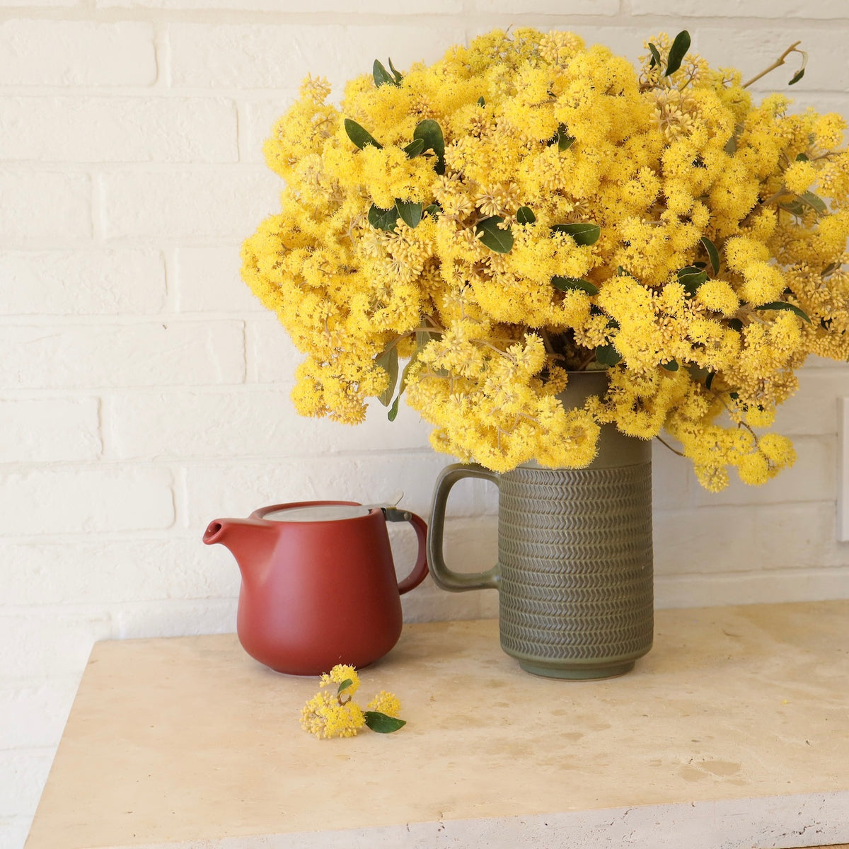 A large bouquet of bright yellow kumarahou flowers in a vase next to e red tea pot