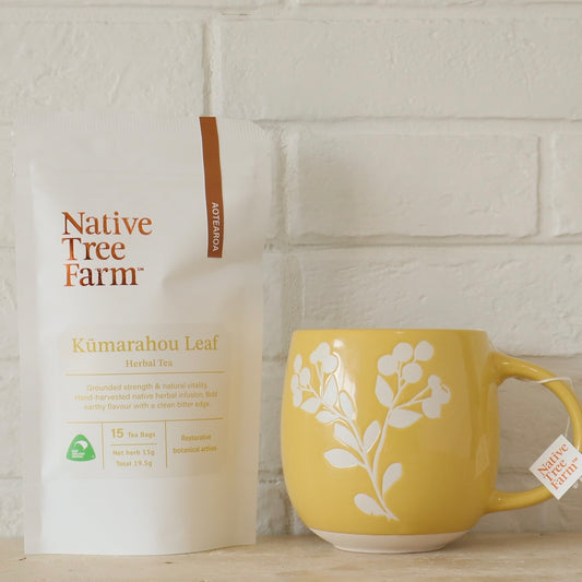 Yellow mug with white floral design next to a 'Native Tree Farm' tea bag on a light brick wall background.