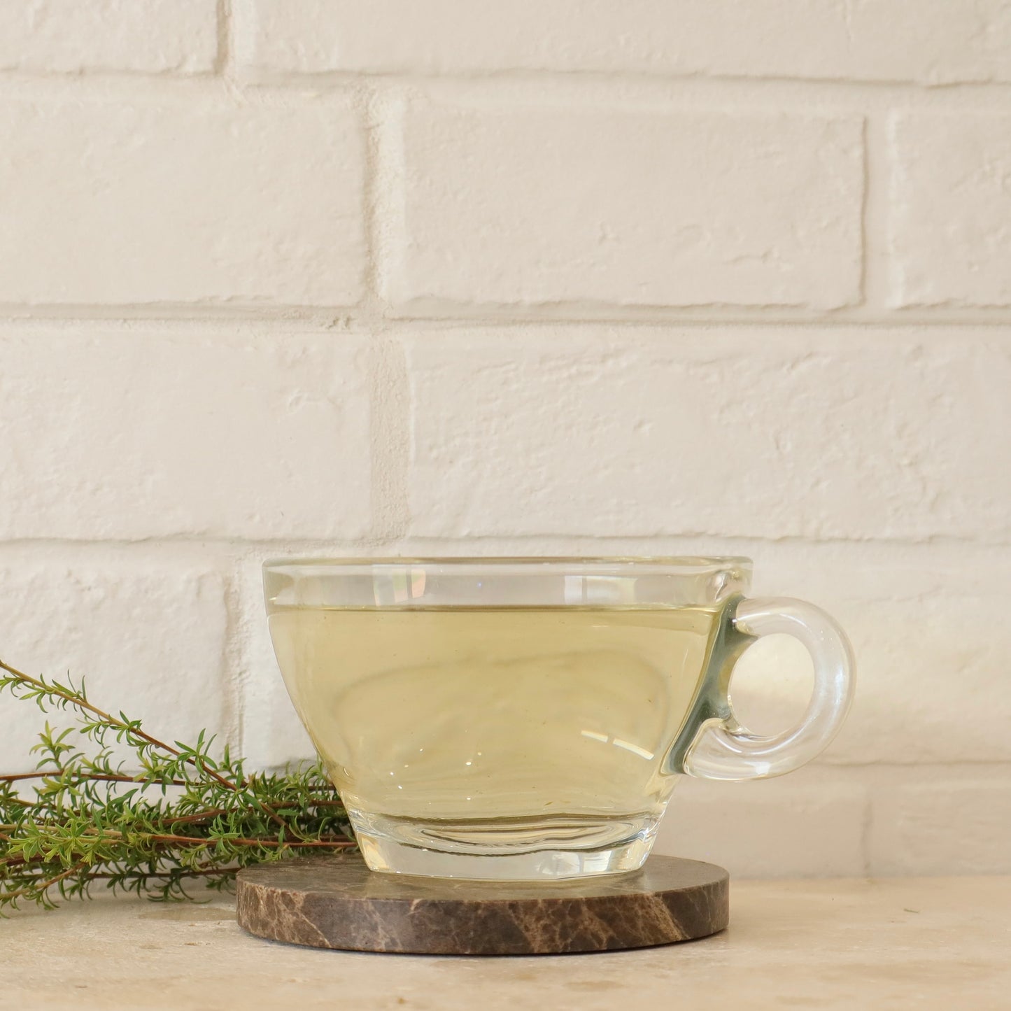 Clear glass mug with manuka calm tea on a wooden coaster against a white brick wall.