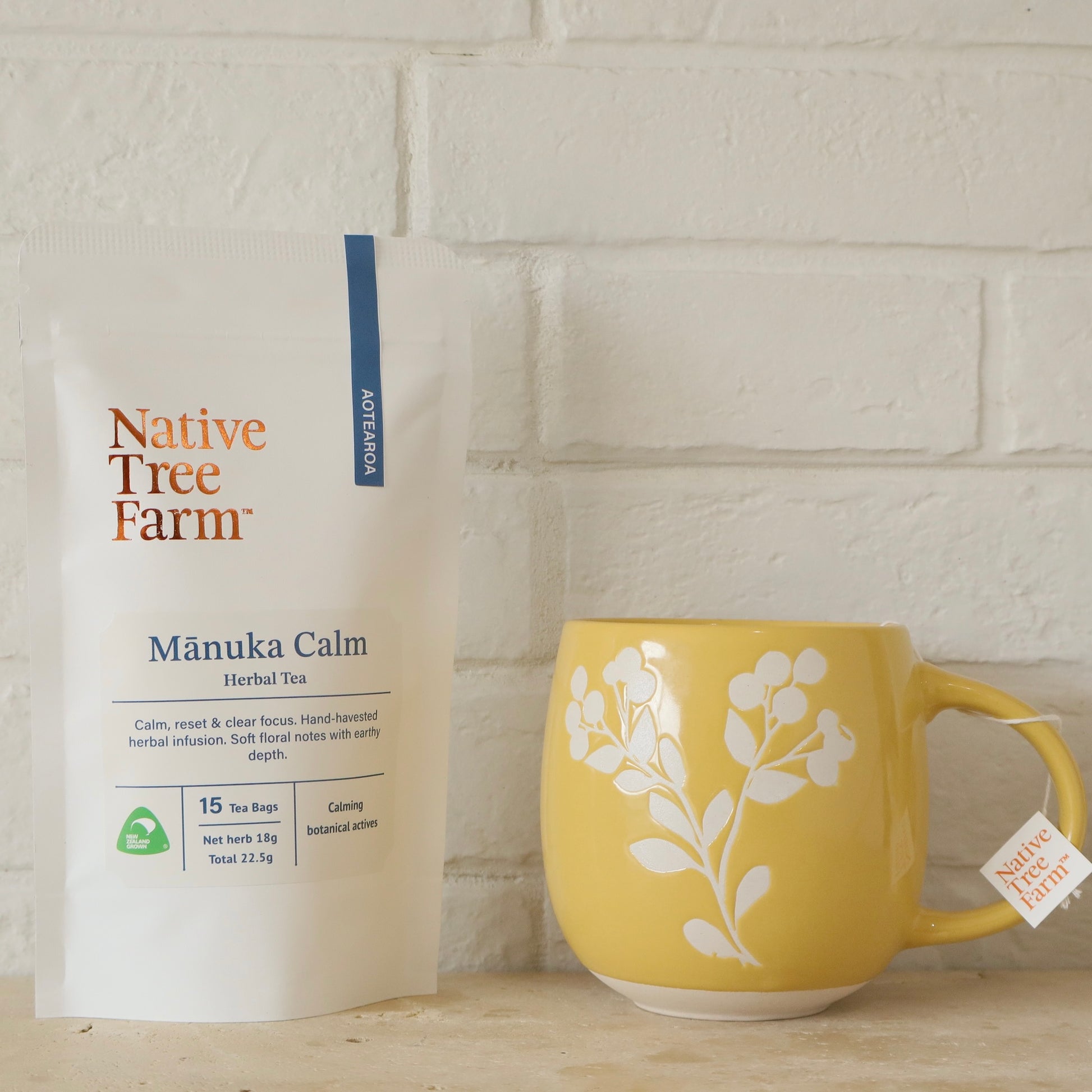 Yellow mug with floral design and Native Tree Farm tea packaging against a white brick wall.