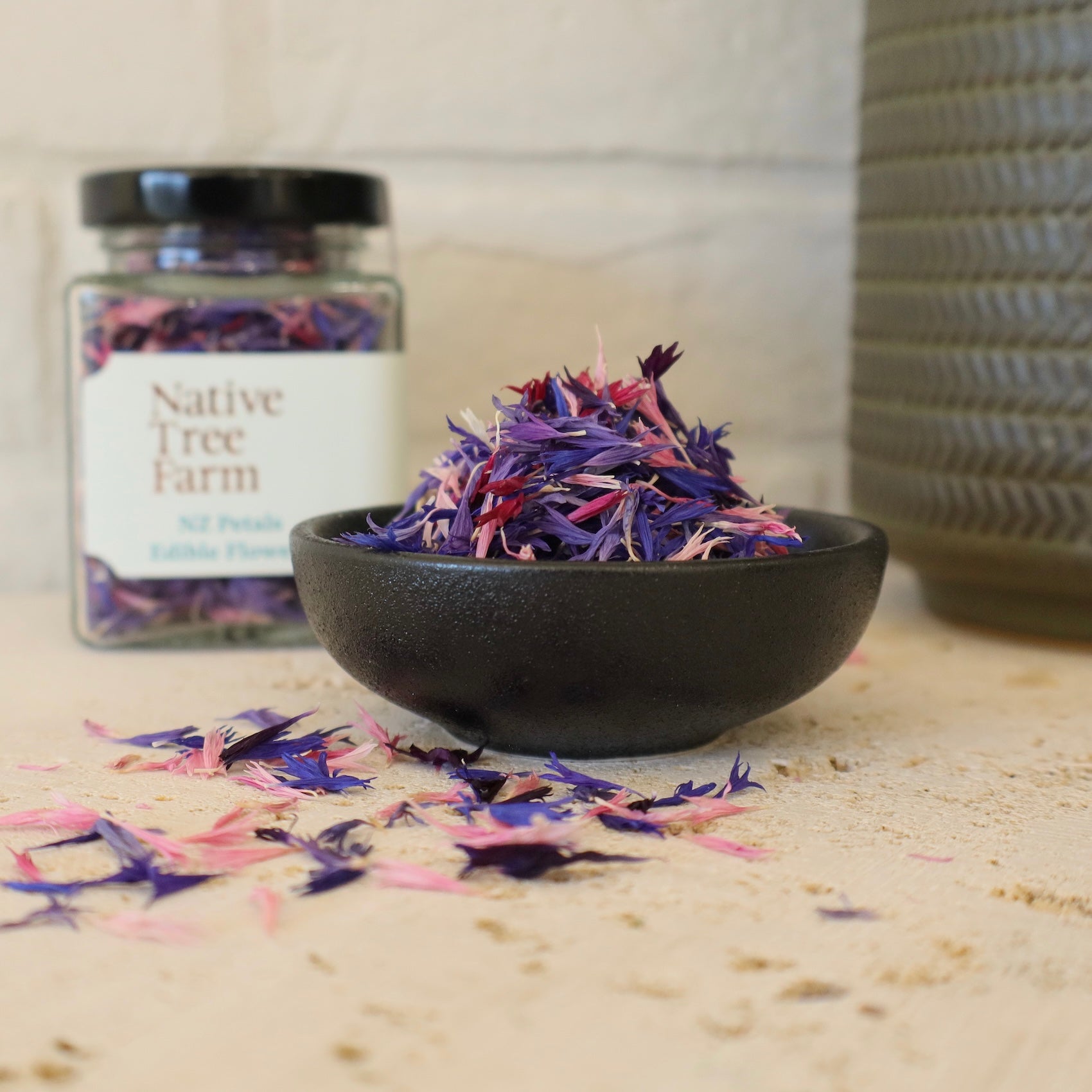 A jar of Native Tree Farm edible flowers with a black bowl next to it filled with the petals.