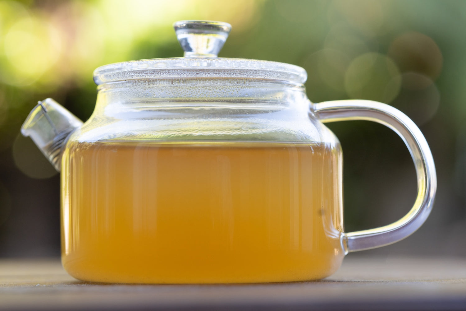 Clear glass teapot with a handle filled with yellow kumarahou tea on a blurred natural background