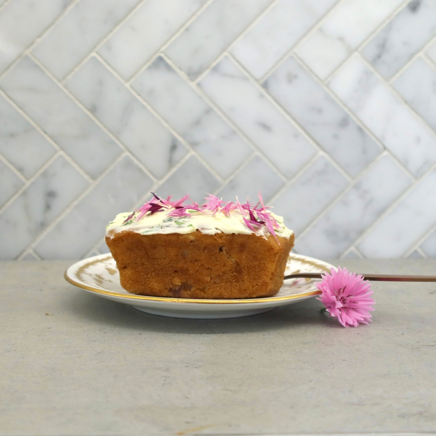 Small loaf cake with icing and decorated with Native Tree Farm, pink organic edible flower petals, sitting on a kitchen table 
