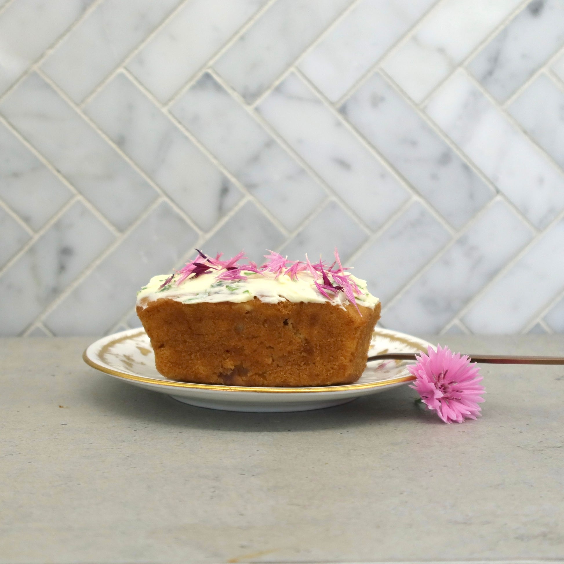 Small loaf cake with icing and decorated with Native Tree Farm, pink organic edible flower petals, sitting on a kitchen table 