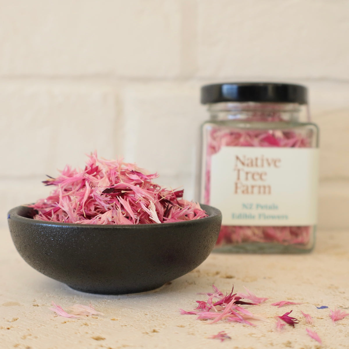 a small black pottle filled with Native Tree Farm pink edible flower petals on a stone kitchen bench top
