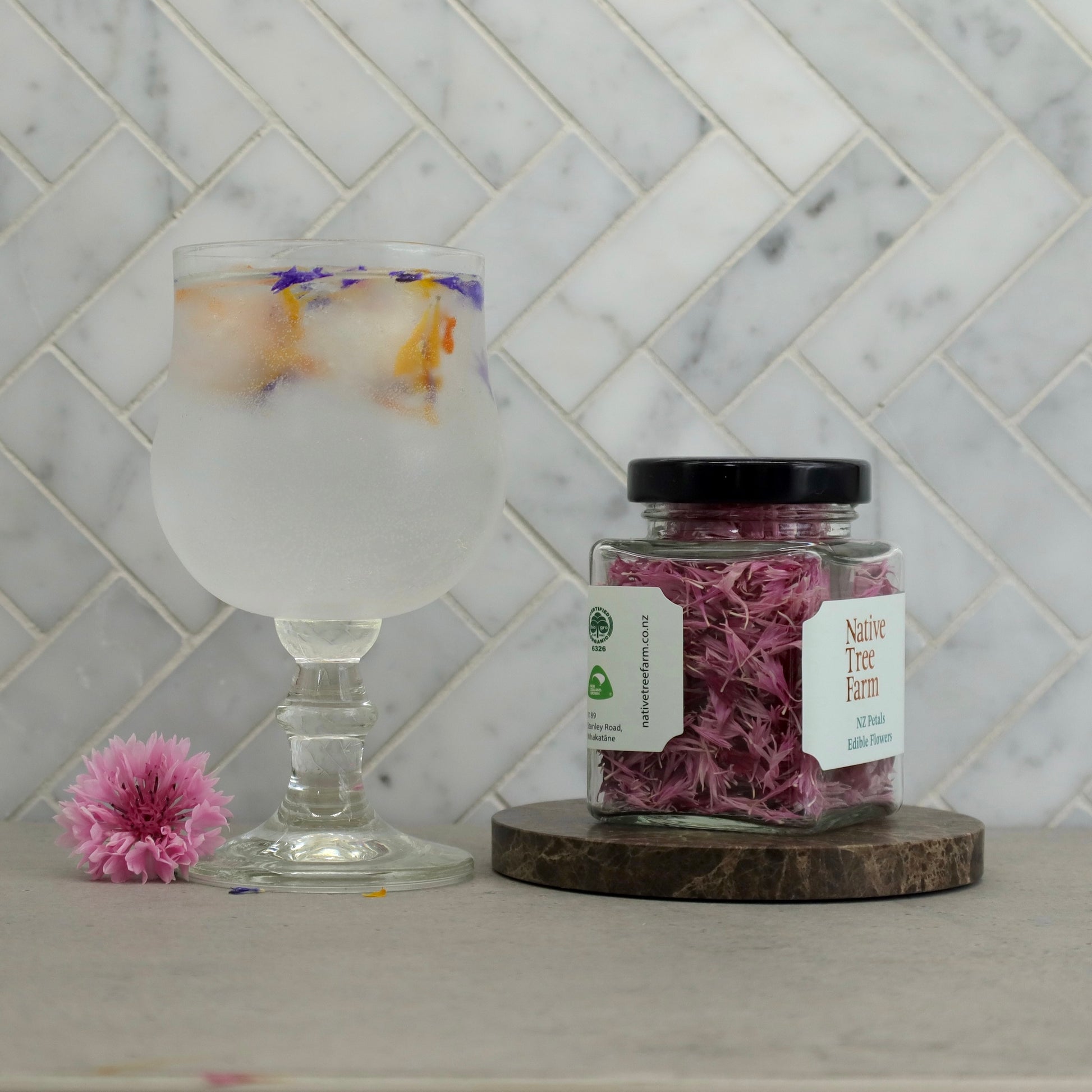 Jar of Native Tree Farm  pink edible flower petals on a stone bench next to a glass of bubbly